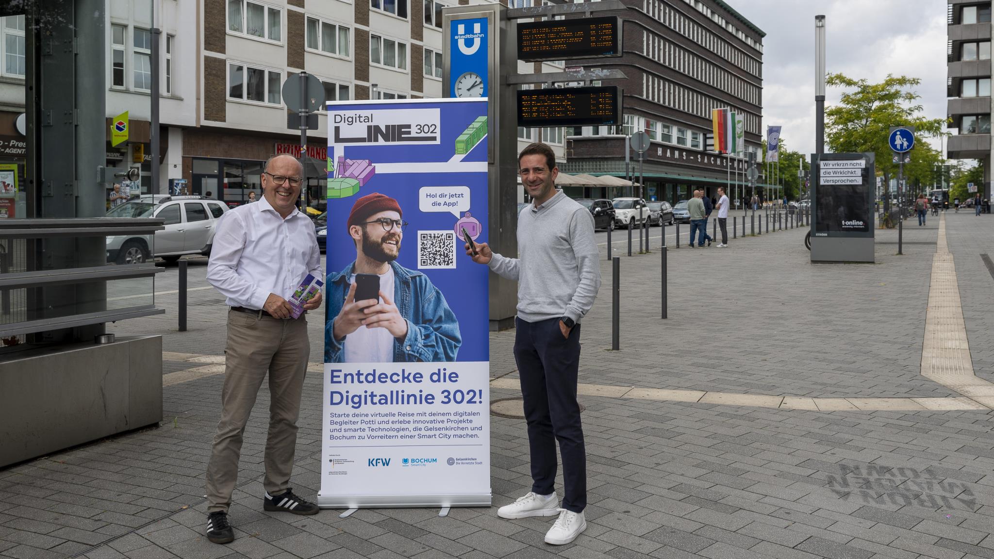 Zwei Männer neben einem Aufsteller, im Hintergrund das Hans-Sachs-Haus