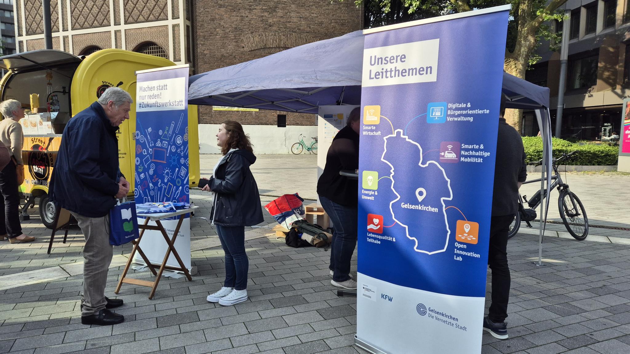 Foto: Stand auf Platz mit Roll-ups und Mann, der sich informiert.