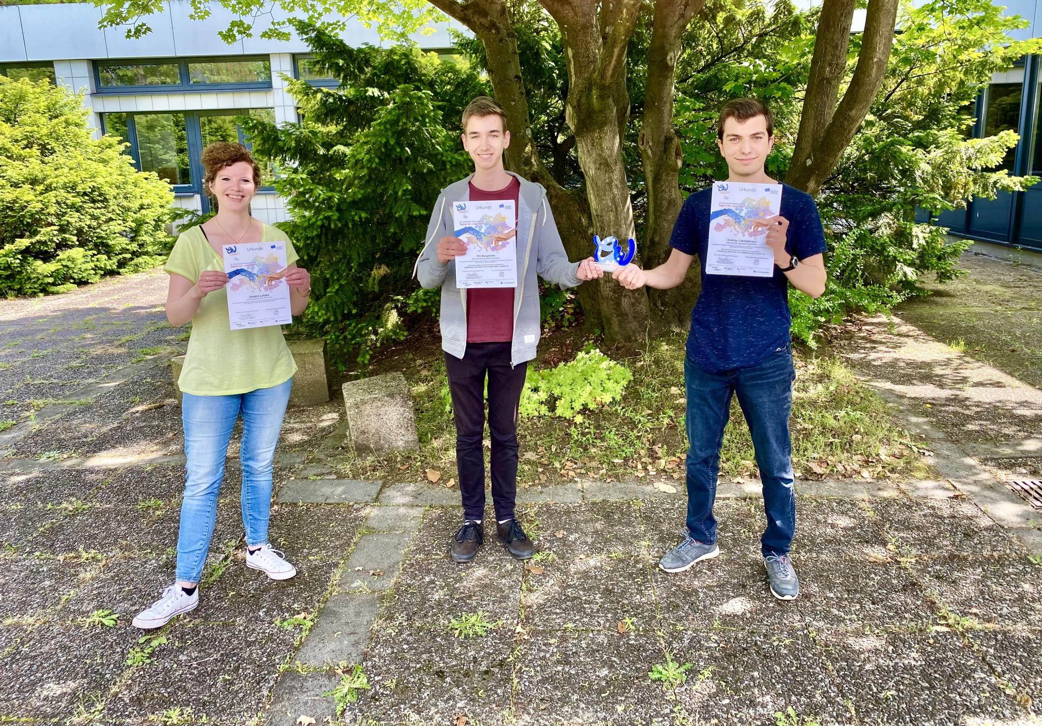 Foto: Drei Personen auf Schulhof halten Urkunden in die Kamera.