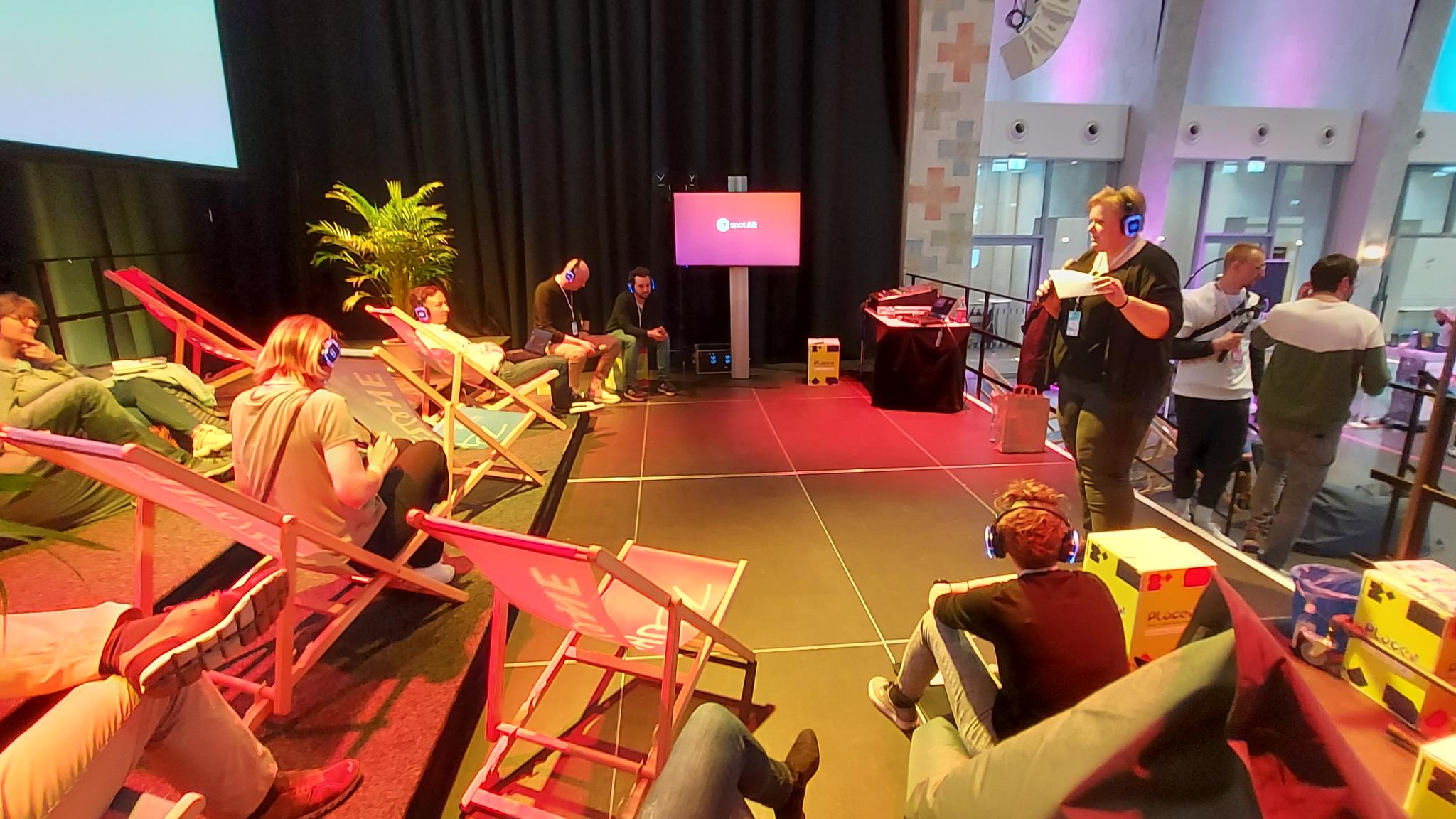 Moderatorin steht auf Bühne; Teilnehmende sitzen auf Liegestühlen.