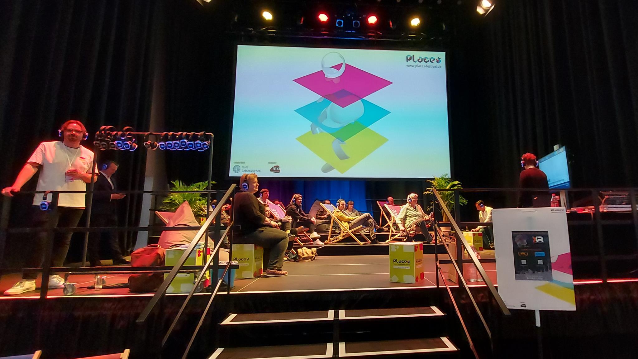 Foto: Bühne der Heilig-Kreuz-Kirche mit großer Leinwand im Hintergrund; auf der Bühne die Network-Area des Places Festivals mit Liegestühlen, Sitzhockern und interessiertem Publikim