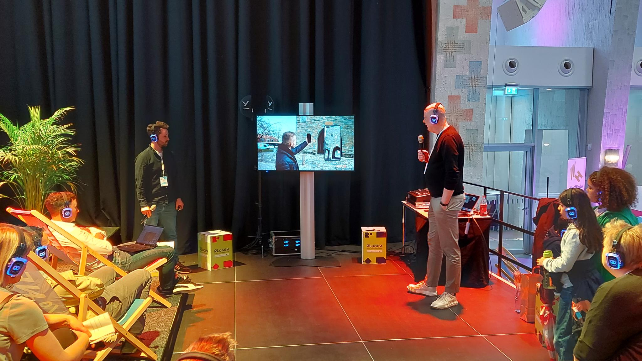 Foto: Zwei Referenten stehen in Network-Area des Places auf der Bühne der Heilig-Kreuz-Kirche und stellen ihr Projekt vor; links und rechts sitzen interessierte Zuschauer in Liegestühlen oder stehen am Rand