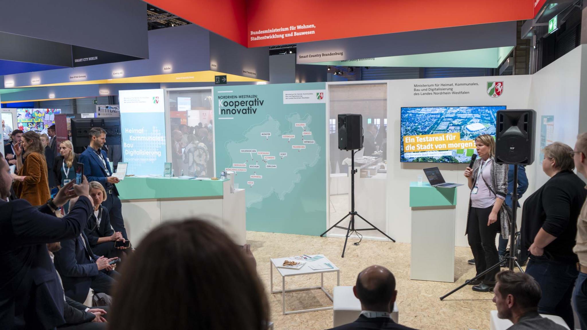 Foto: Speakers Corner auf dem Messestand des MHKBD, vorne zuhörende Menschen, im Hintergrund Standelemente, Bildschirm und Referentin