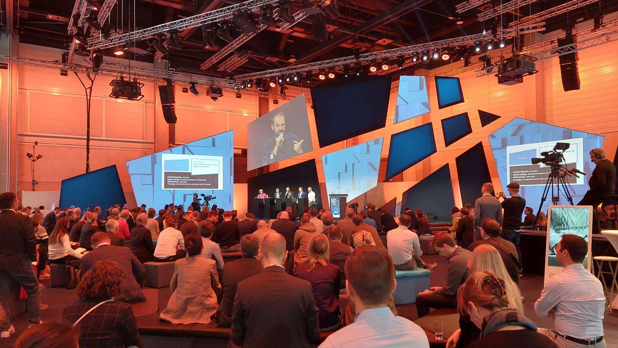 Publikum sitzt vor einer Bühne, auf der ein Redner einen Vortrag hält.